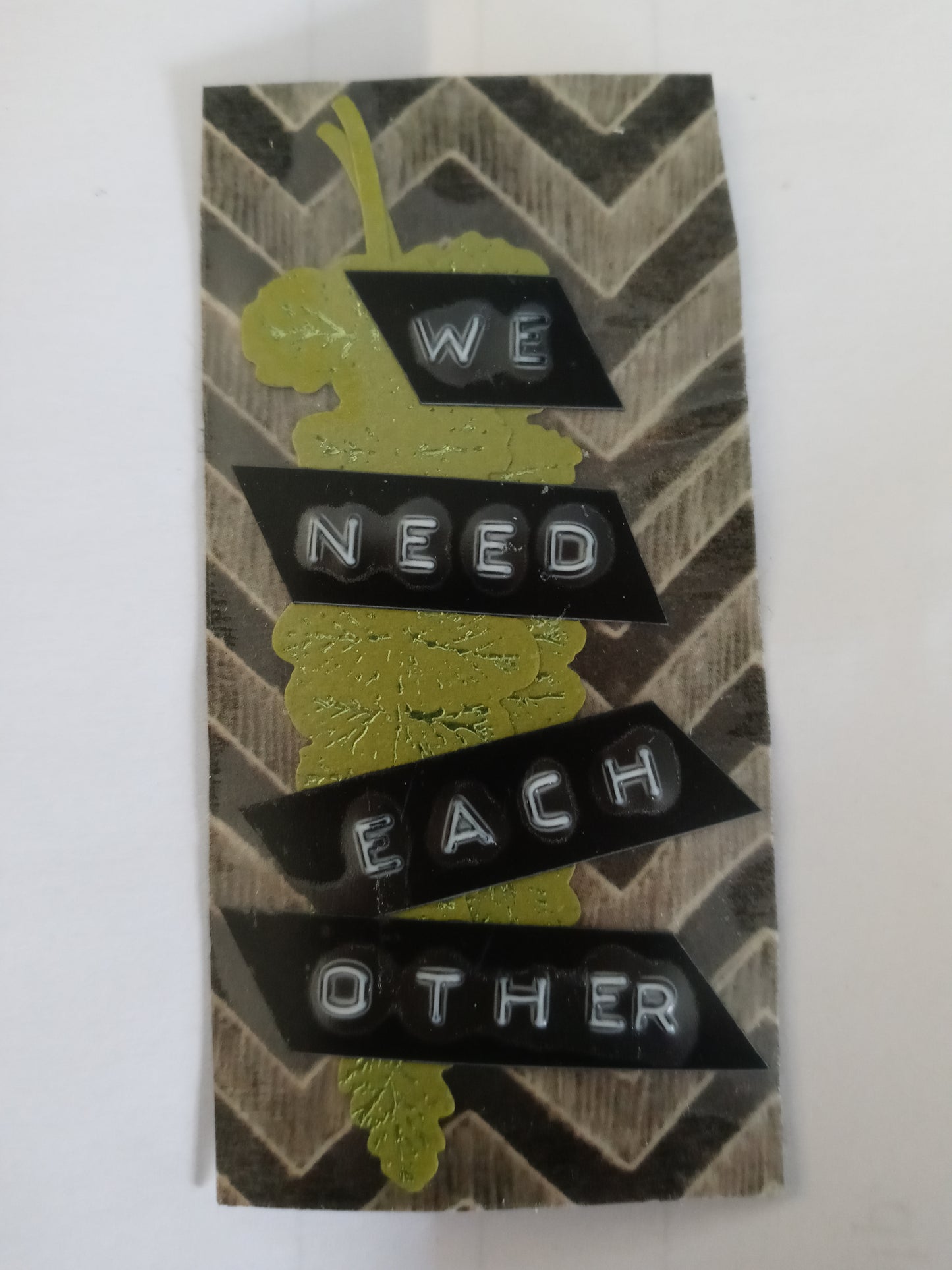 A small, long rectangular sticker with a background of black and beige zig zags, with a shiny green diecut leaf and the words "WE NEED EACH OTHER" printed from a label maker on top.