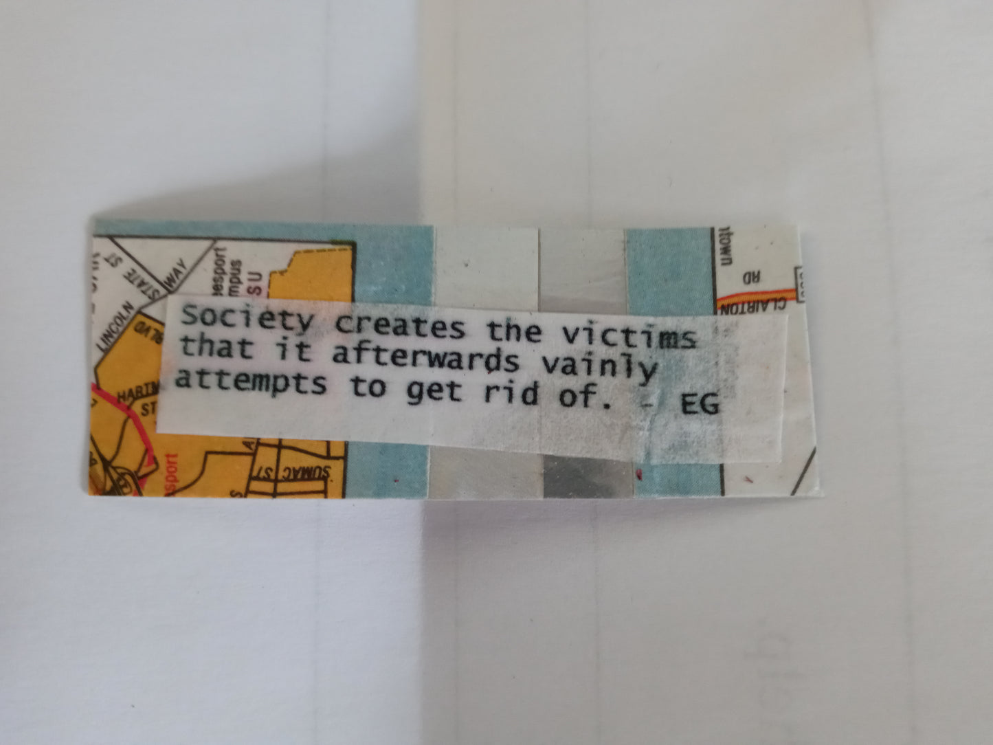 A small, rectangular handmade sticker with a map background and a quote printed on white paper that reads "Society creates the victims that it afterwards vainly attempts to get rid of. - EG"