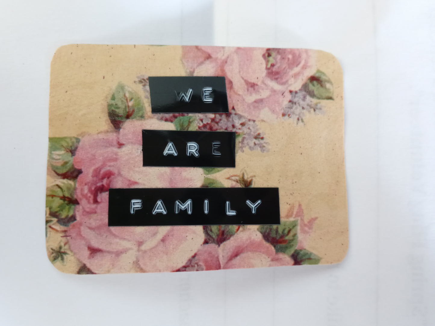 A handmade sticker in the shape of an irregular, rounded off rectangle with a floral background in beige, pink and green. Over top there are 3 lines of text made with a label maker that read "WE ARE FAMILY"