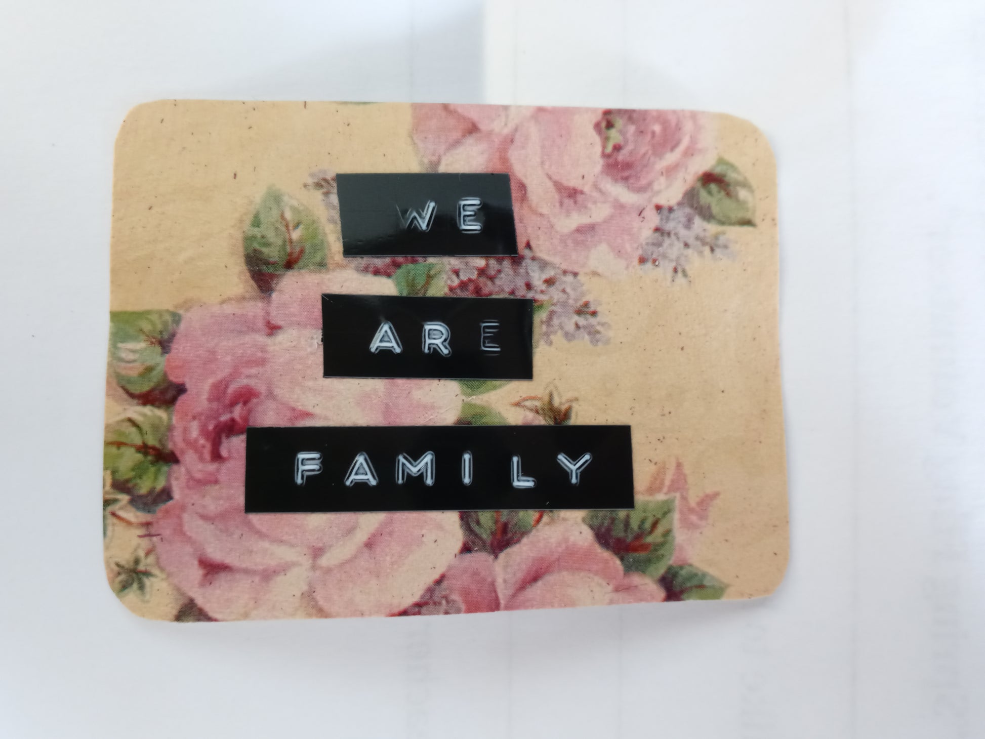 A handmade sticker in the shape of an irregular, rounded off rectangle with a floral background in beige, pink and green. Over top there are 3 lines of text made with a label maker that read "WE ARE FAMILY"