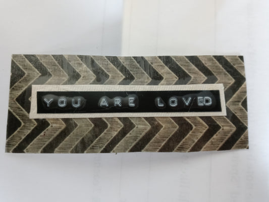Handmade sticker with "YOU ARE LOVED" printed from a label maker centered on a beige rectangle, centered on an uneven black and beige zig zag patterned background rectangle.