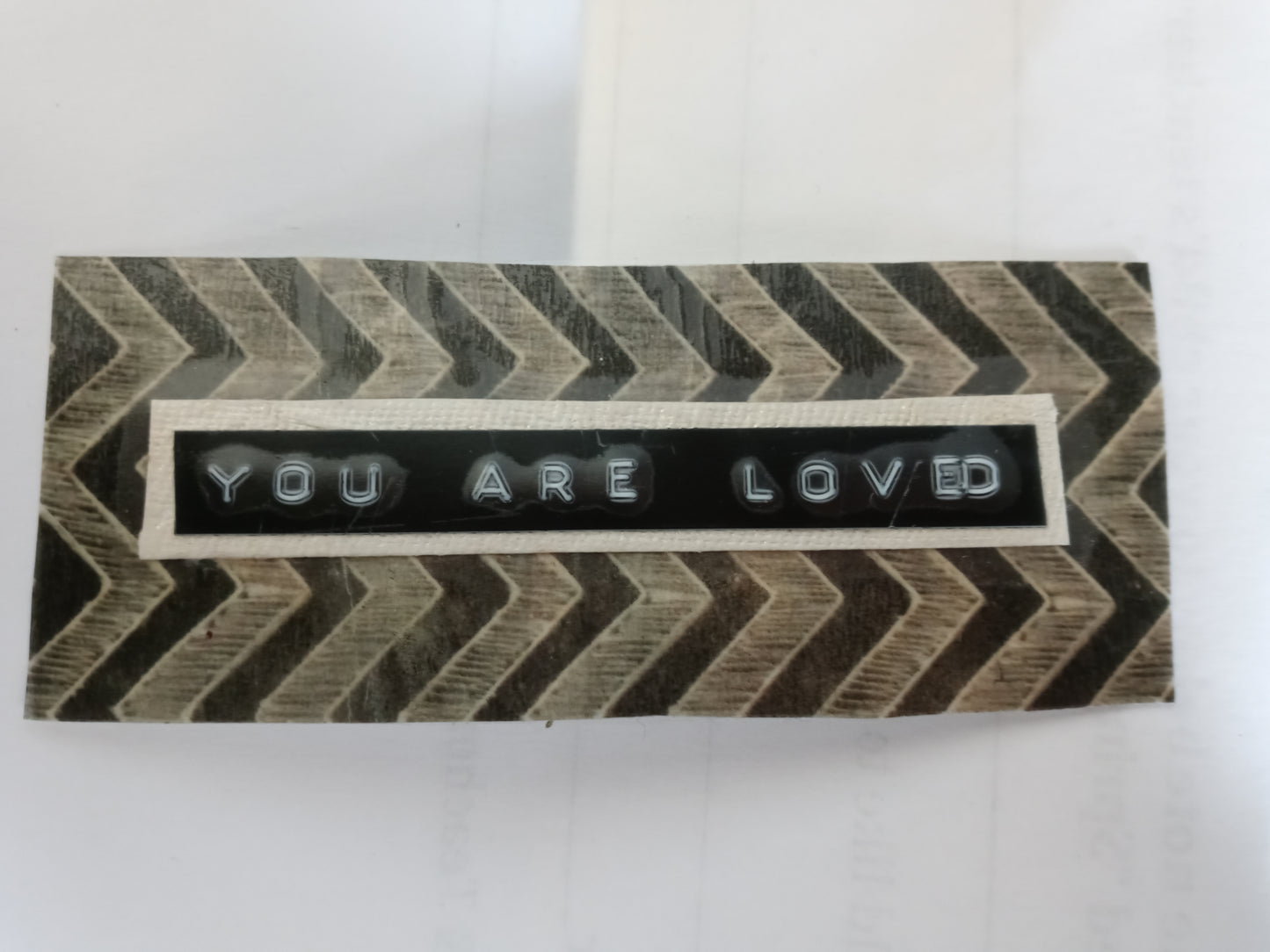 Handmade sticker with "YOU ARE LOVED" printed from a label maker centered on a beige rectangle, centered on an uneven black and beige zig zag patterned background rectangle.